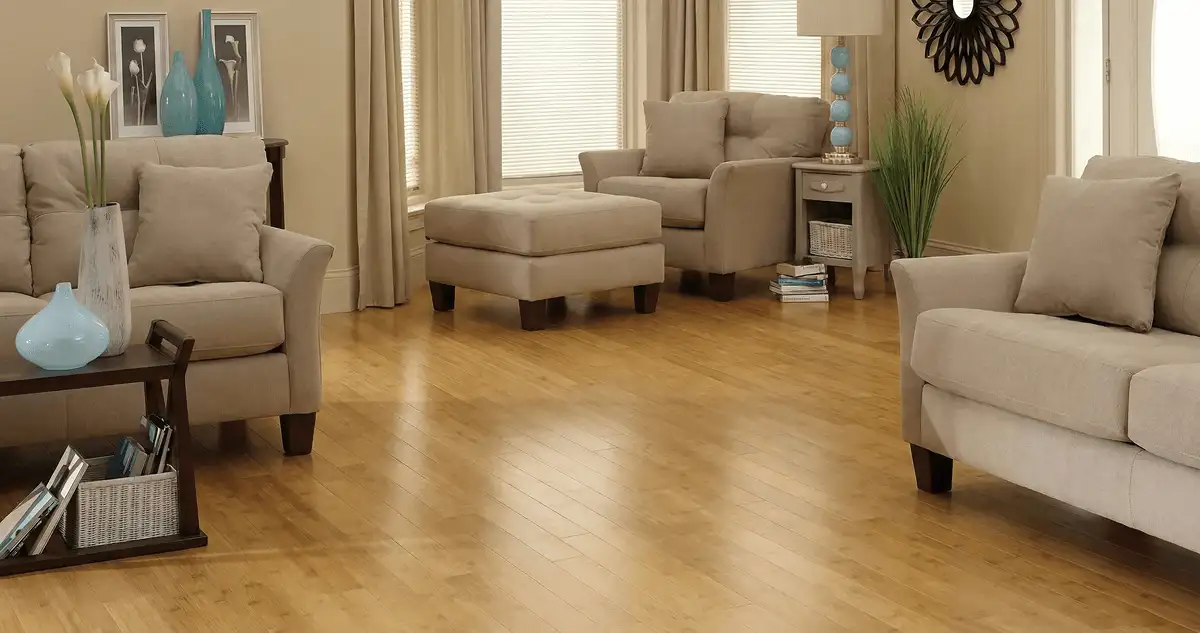 A living room with professionally installed bamboo flooring, beige sofas, a matching ottoman, a side table with a blue lamp, vases, framed pictures, and large windows with blinds and curtains in Bradenton, FL